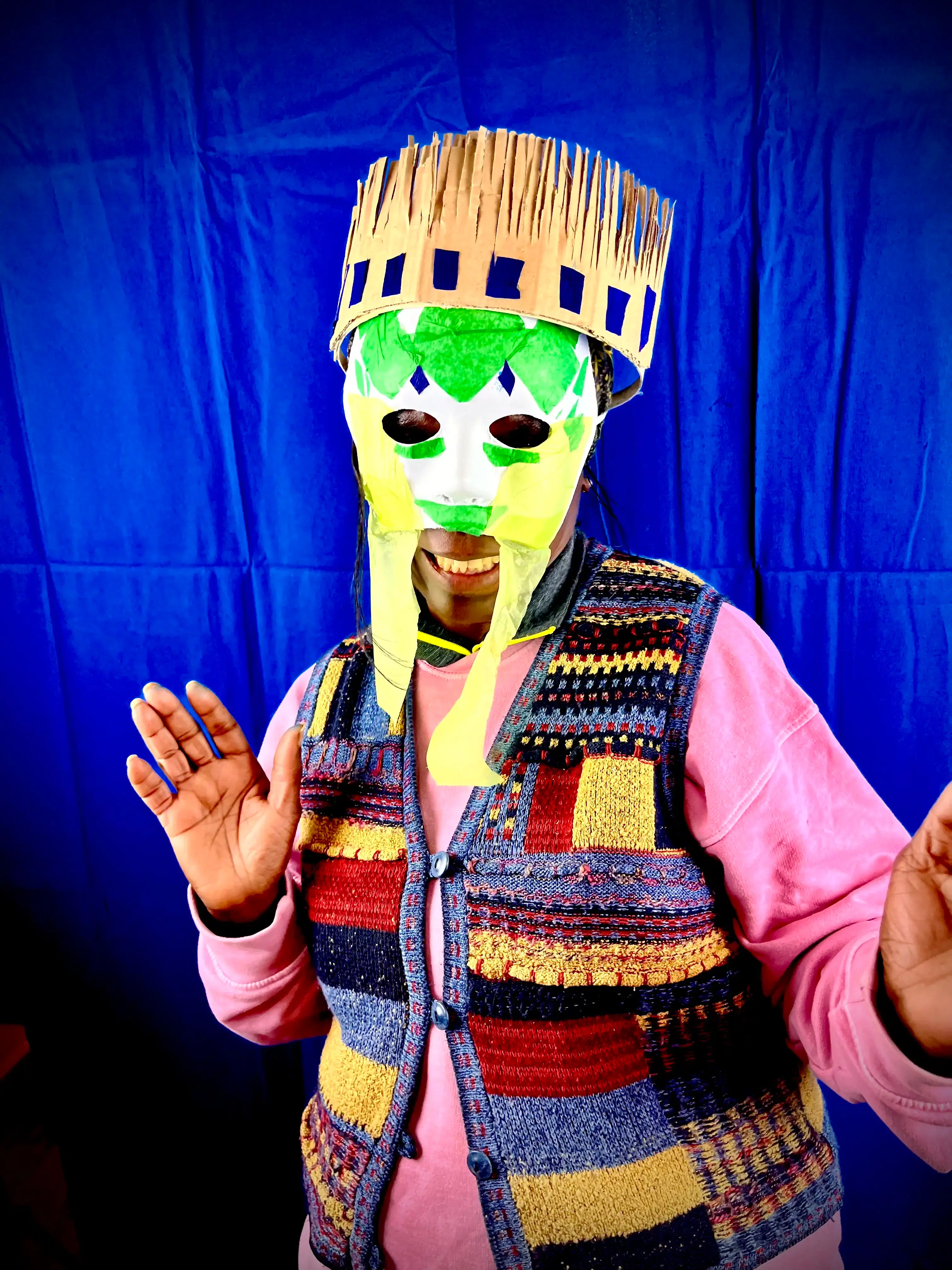 A person wearing a colorful knit vest and a pink shirt, posing cheerfully with their hands up, sporting a creative mask made of paper and cardboard with a playful straw-like top.