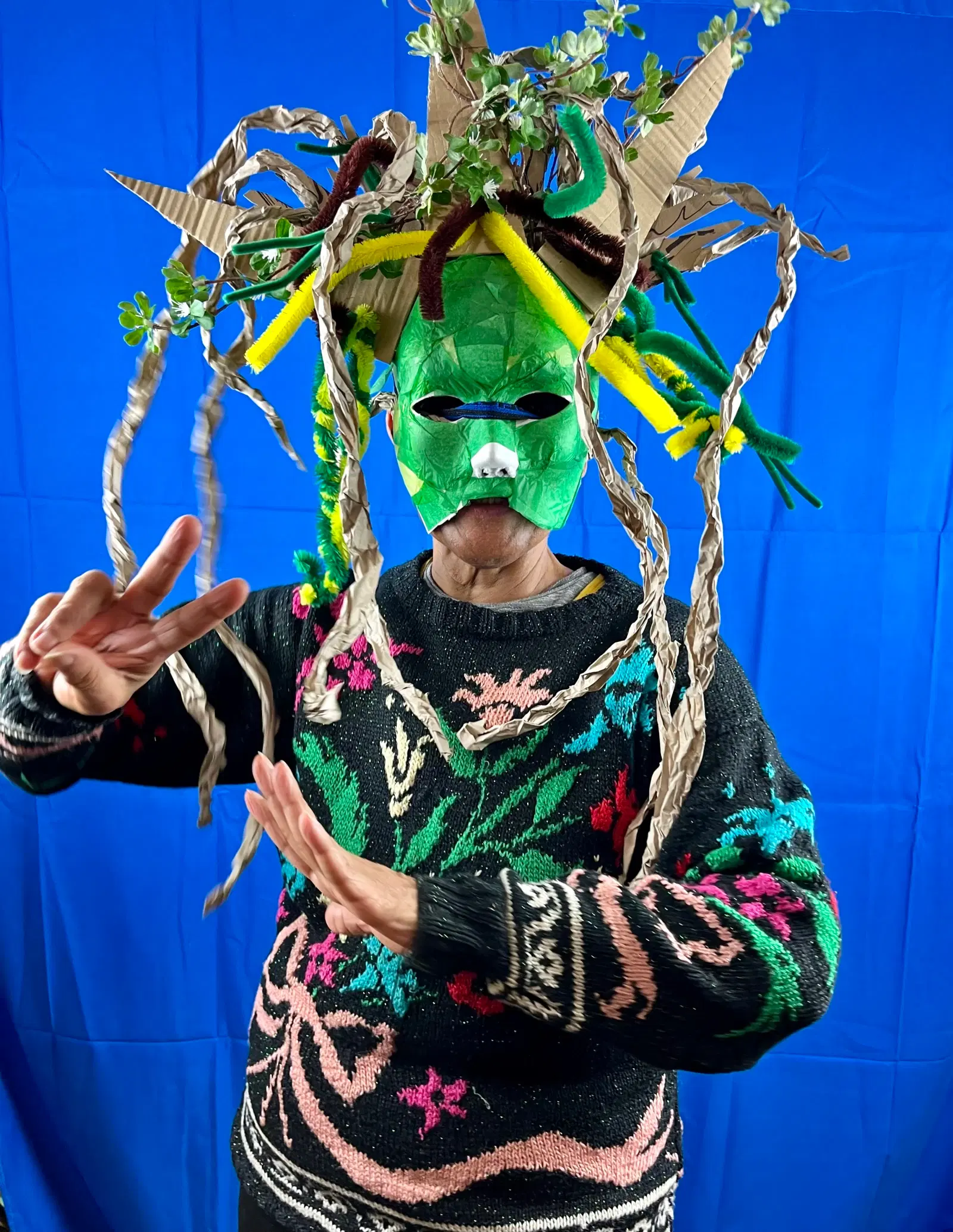 A person in a creative and colorful handmade mask with twisted paper elements and vibrant green tones, posing energetically against a blue backdrop.
