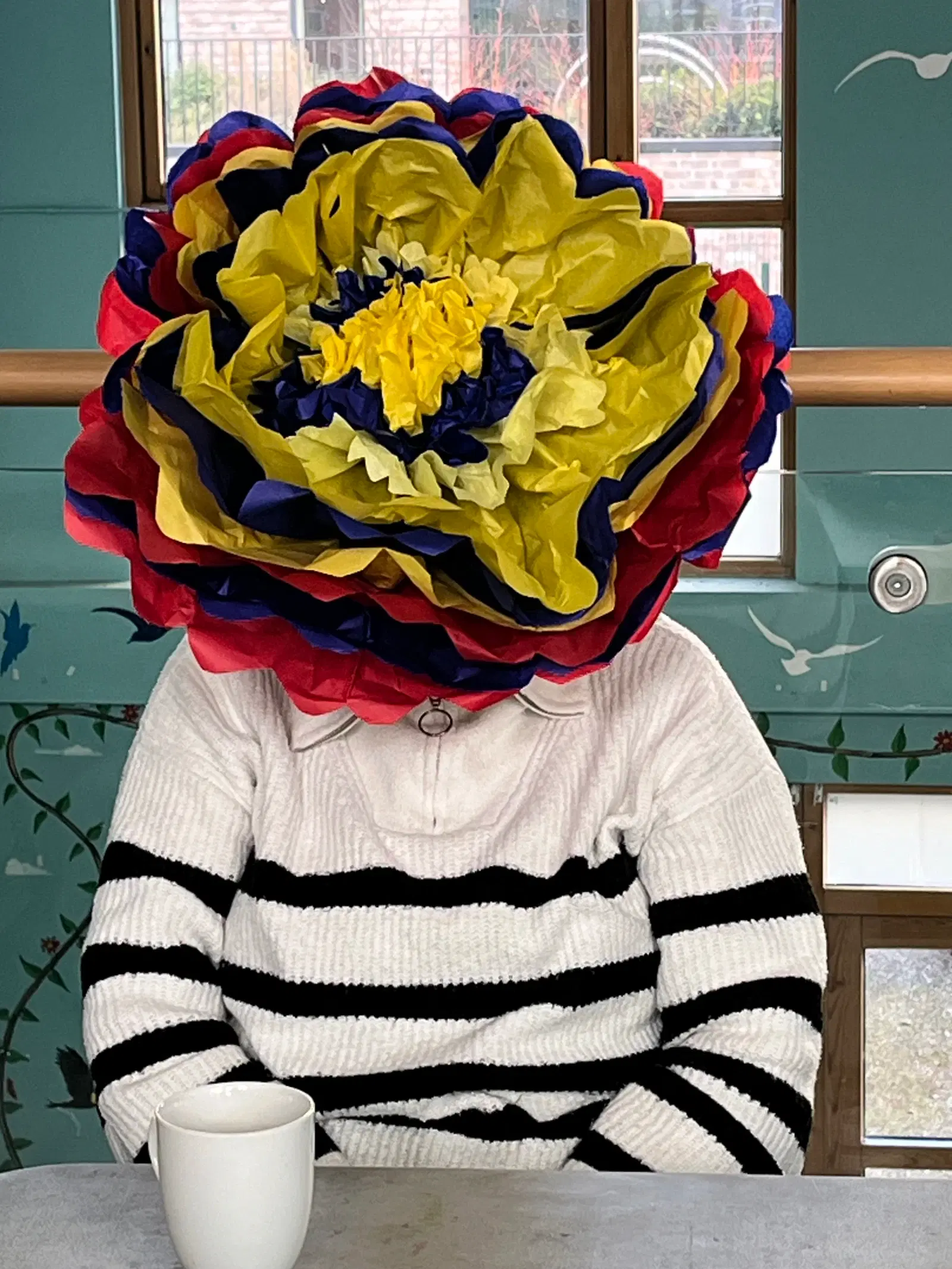 A person seated at a table with an oversized colorful paper flower covering their face, holding a white mug, against a background of windows with garden views.