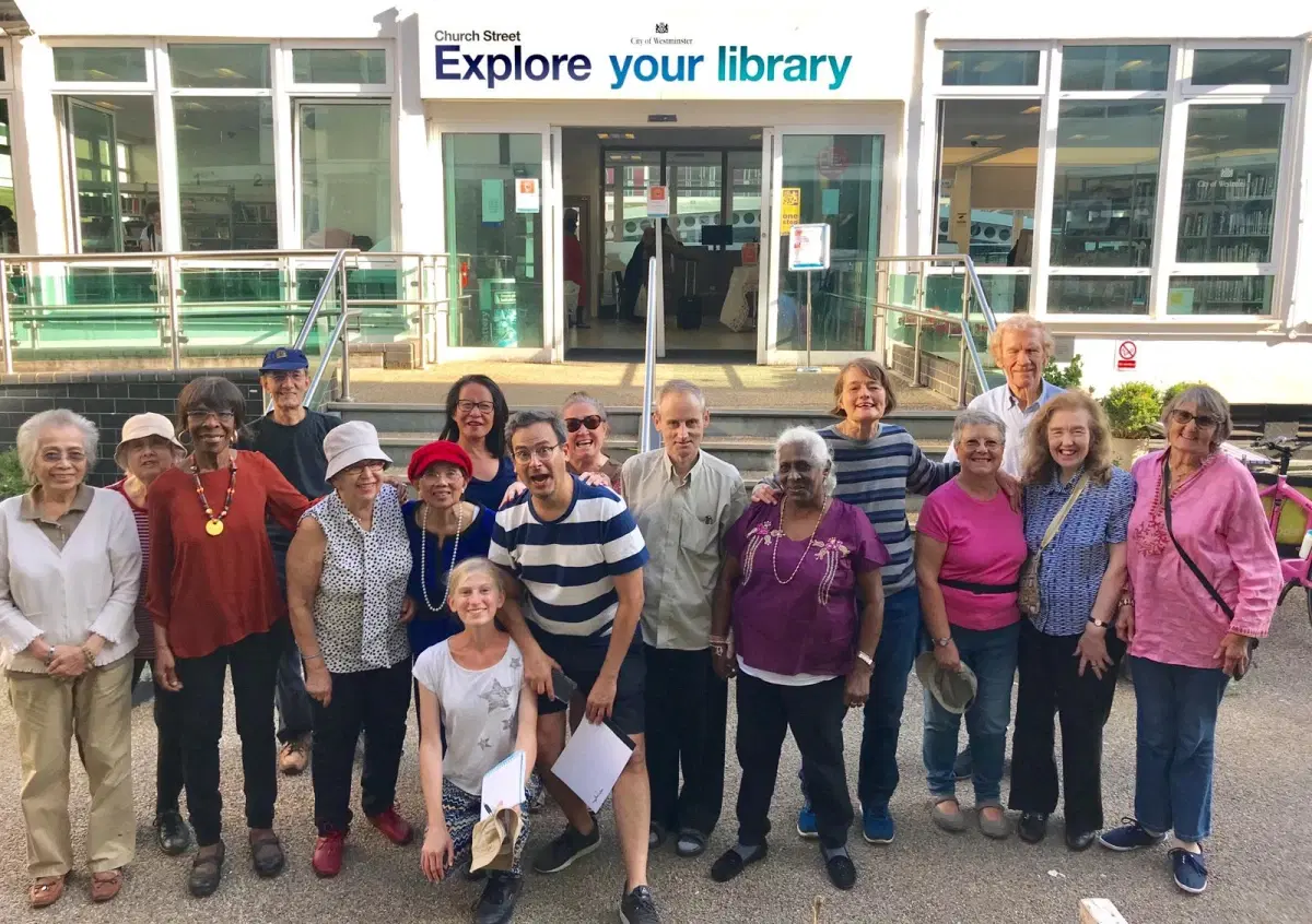 A diverse group of smiling people gathered in front of a library, under a sign that invites passersby to "explore your library," showcasing a sense of community and a love for reading and learning.