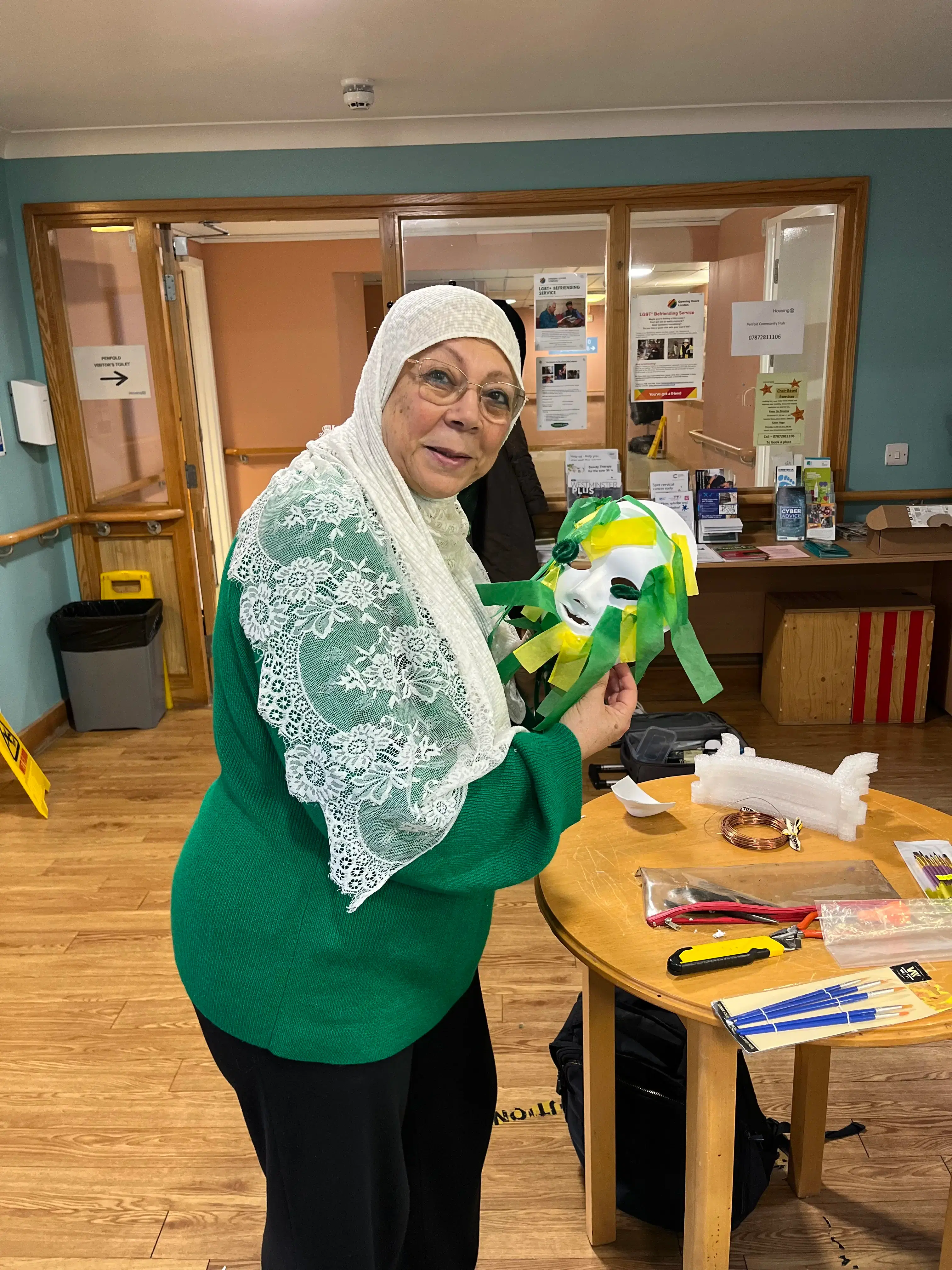 A smiling woman in a green sweater and white lace headscarf holding a colorful handcrafted item, standing in a room with informational flyers on the wall and various items on the table.