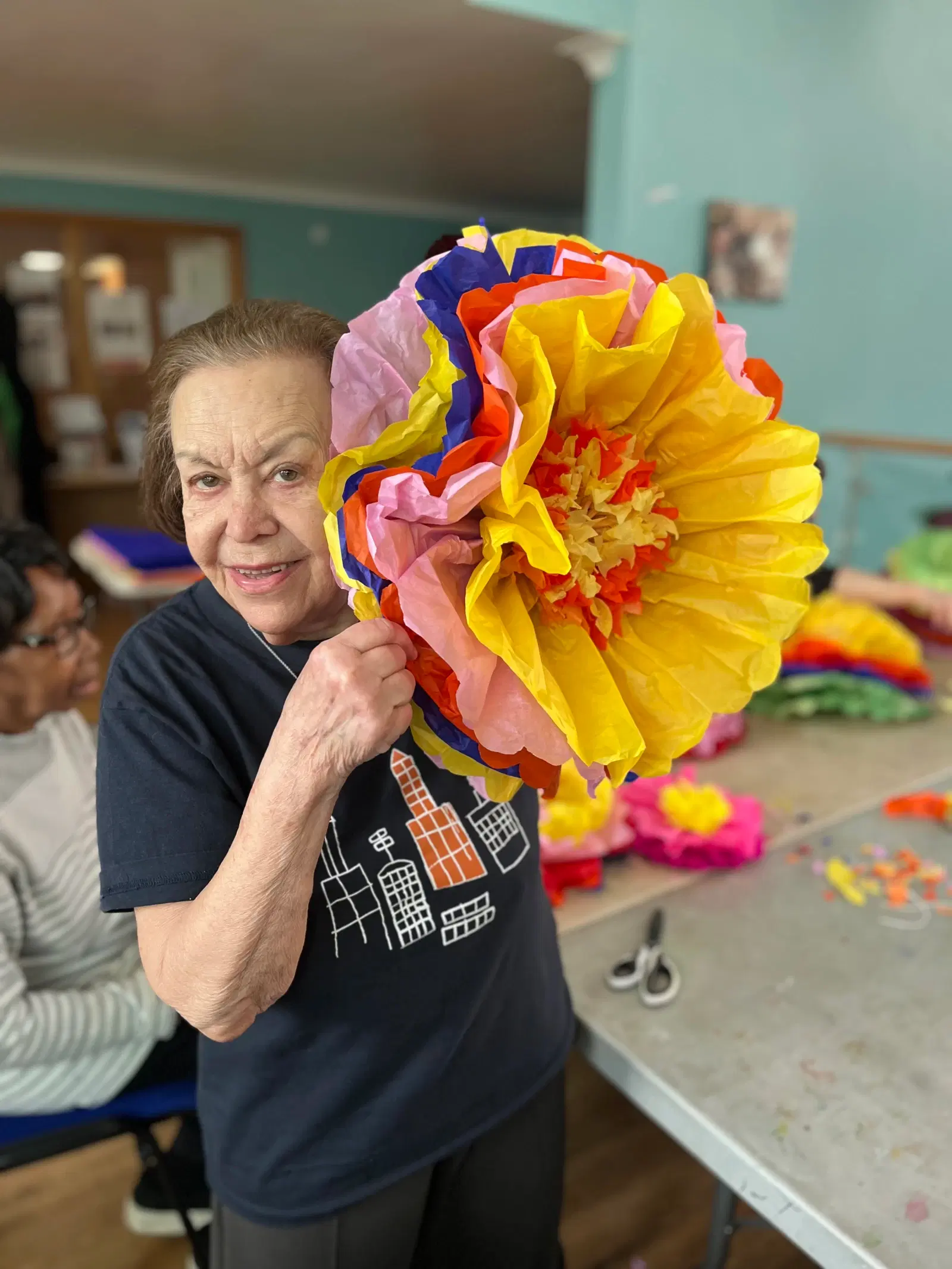 A woman proudly displaying a large, colorful paper flower she has crafted, with a background of a craft-making session in progress.