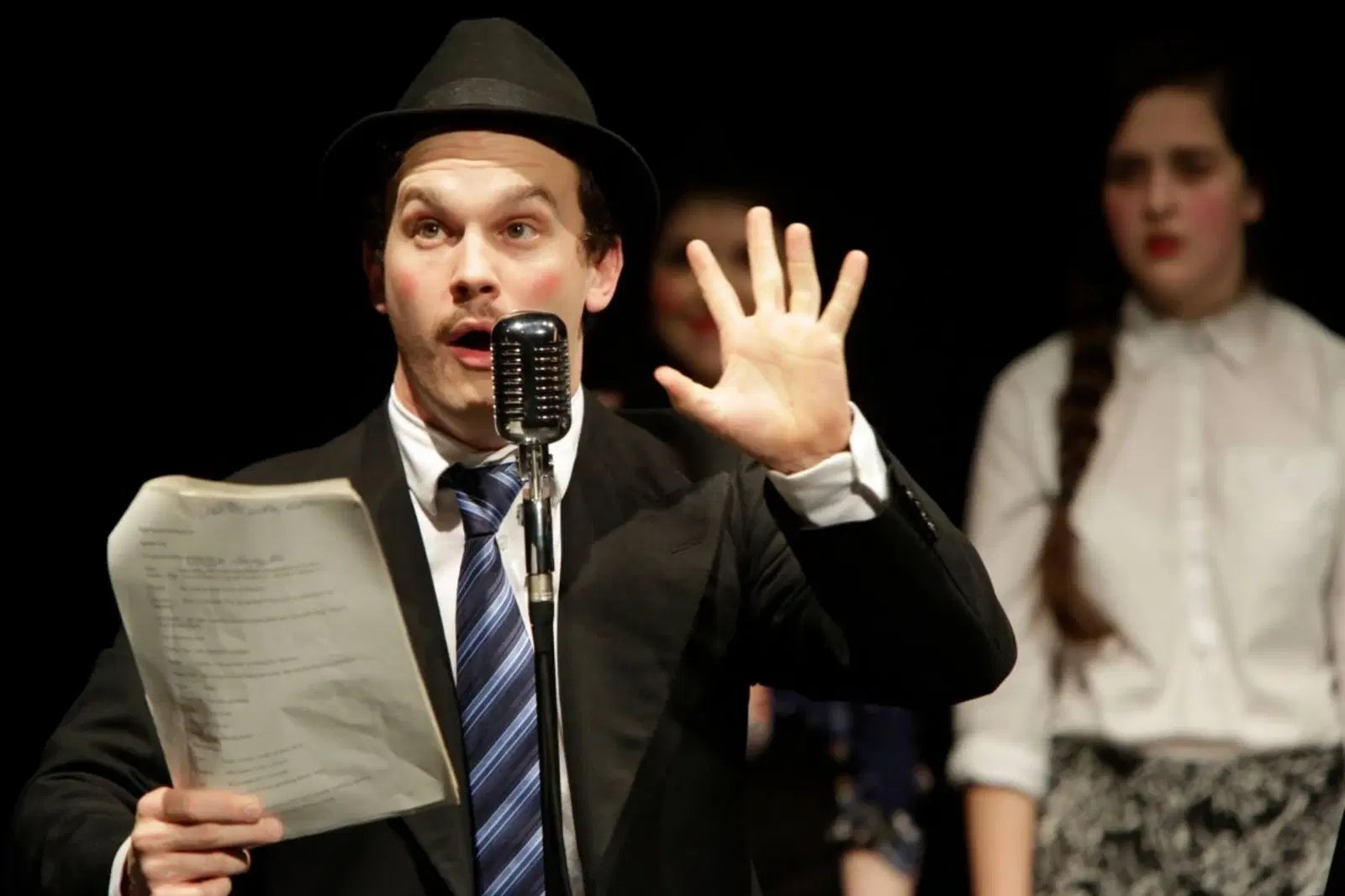A performer in a black hat and suit gestures dramatically into a microphone while holding a script, with a focused actress in the background.