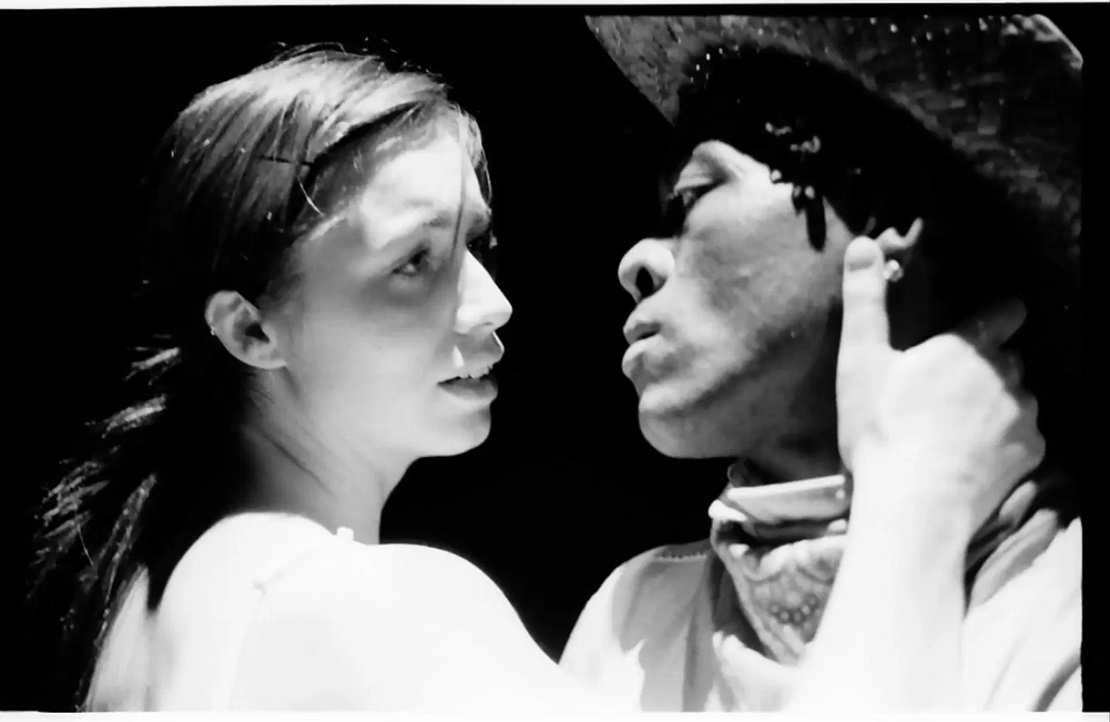 Intense black and white scene of a man and a woman gazing into each other's eyes, conveying a moment of deep connection or confrontation. the man wears a hat and bandana, suggesting a costume, while the woman's bare shoulders and earnest expression add to the dramatic atmosphere.