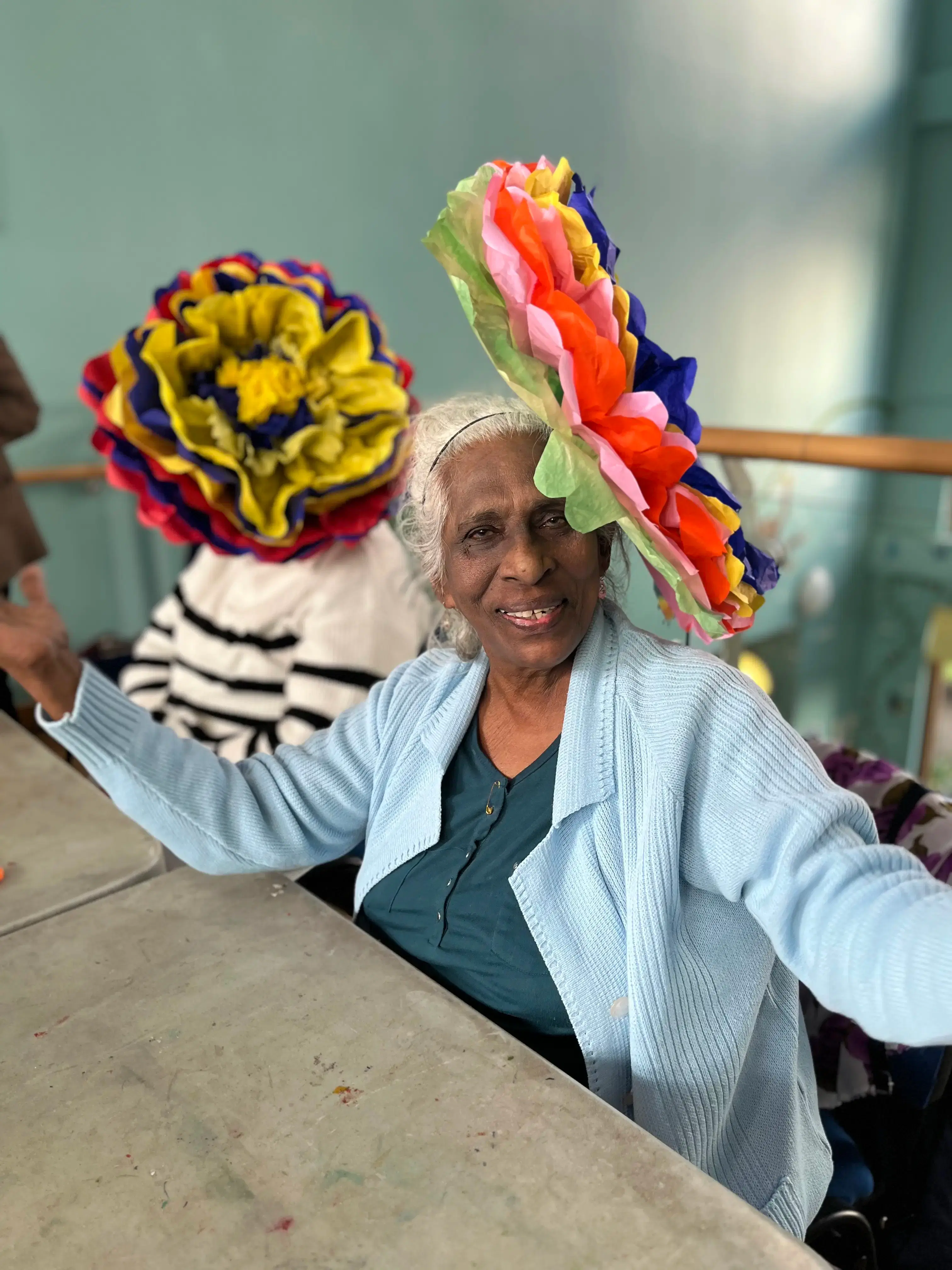 A joyful elderly woman with a vibrant, multicolored paper flower hat, smiling warmly and posing for a picture.