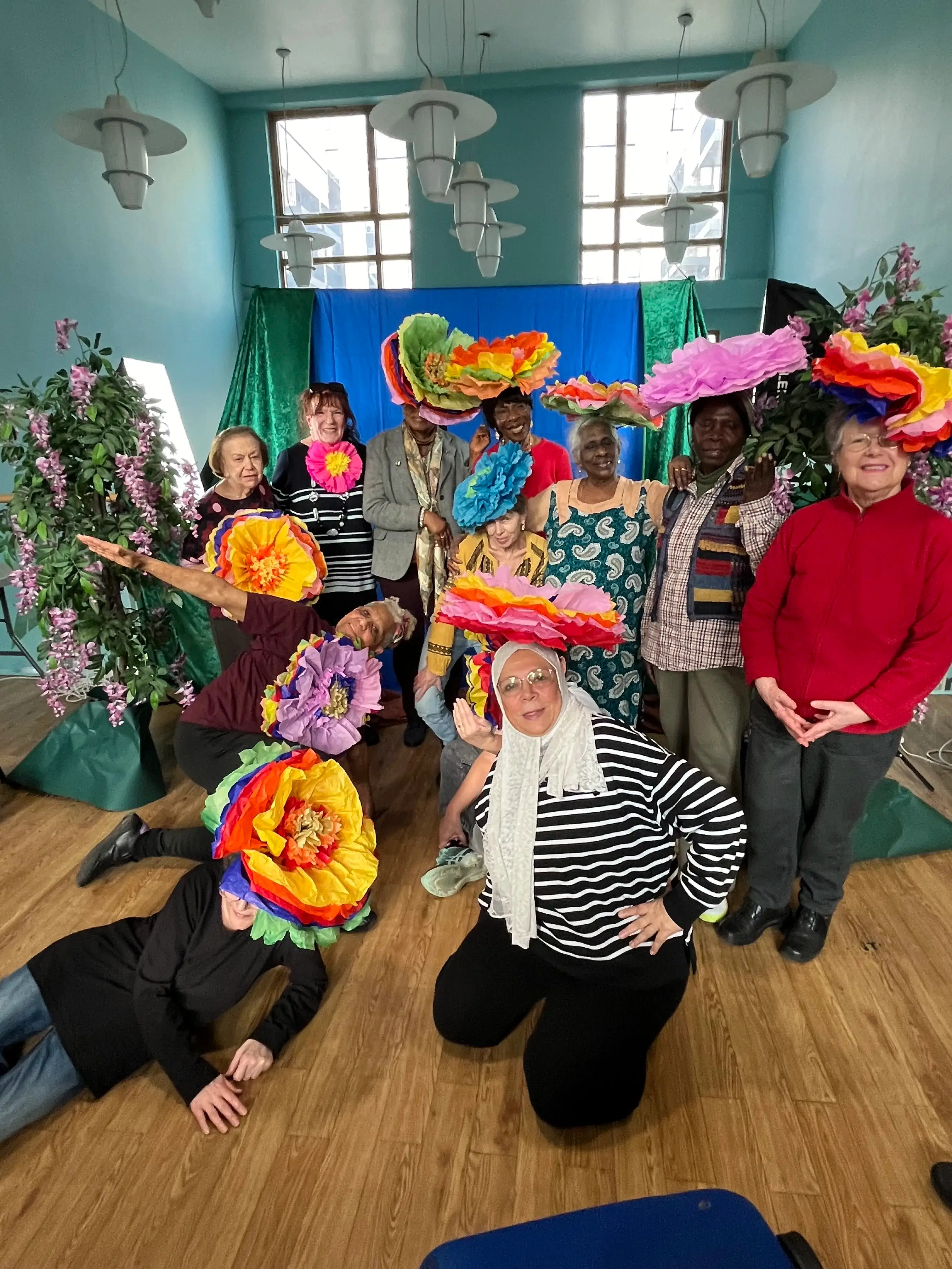 A lively group of people wearing oversized, colorful paper flowers on their heads, posing for a fun and vibrant photo in a room decorated with greenery and festive backdrops.