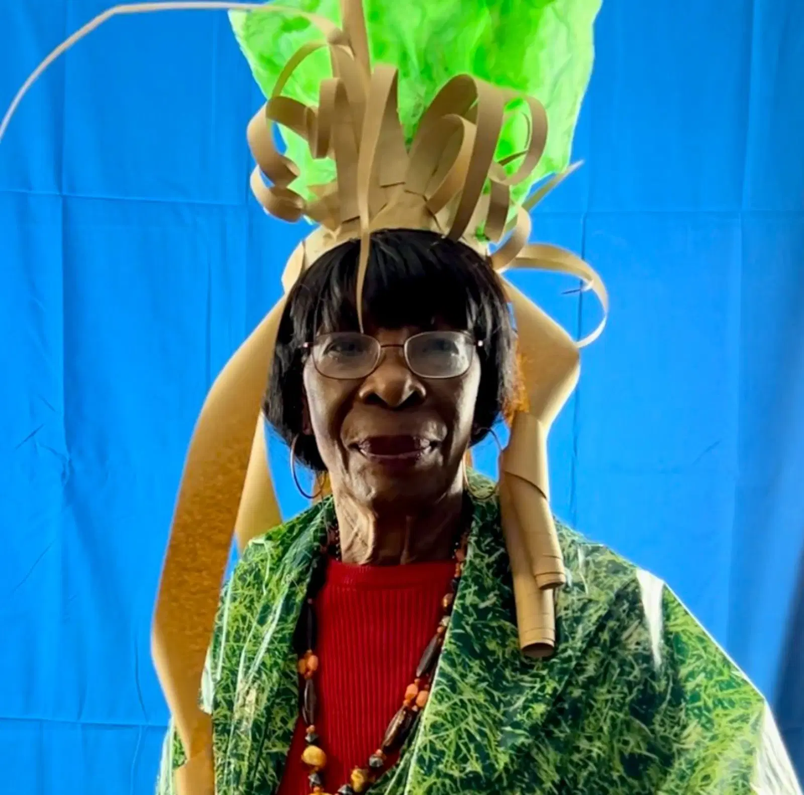A joyful person wearing a whimsical headpiece made of cardboard and paper crafted to look like foliage and plants, with a bright blue background setting off their cheerful demeanor.