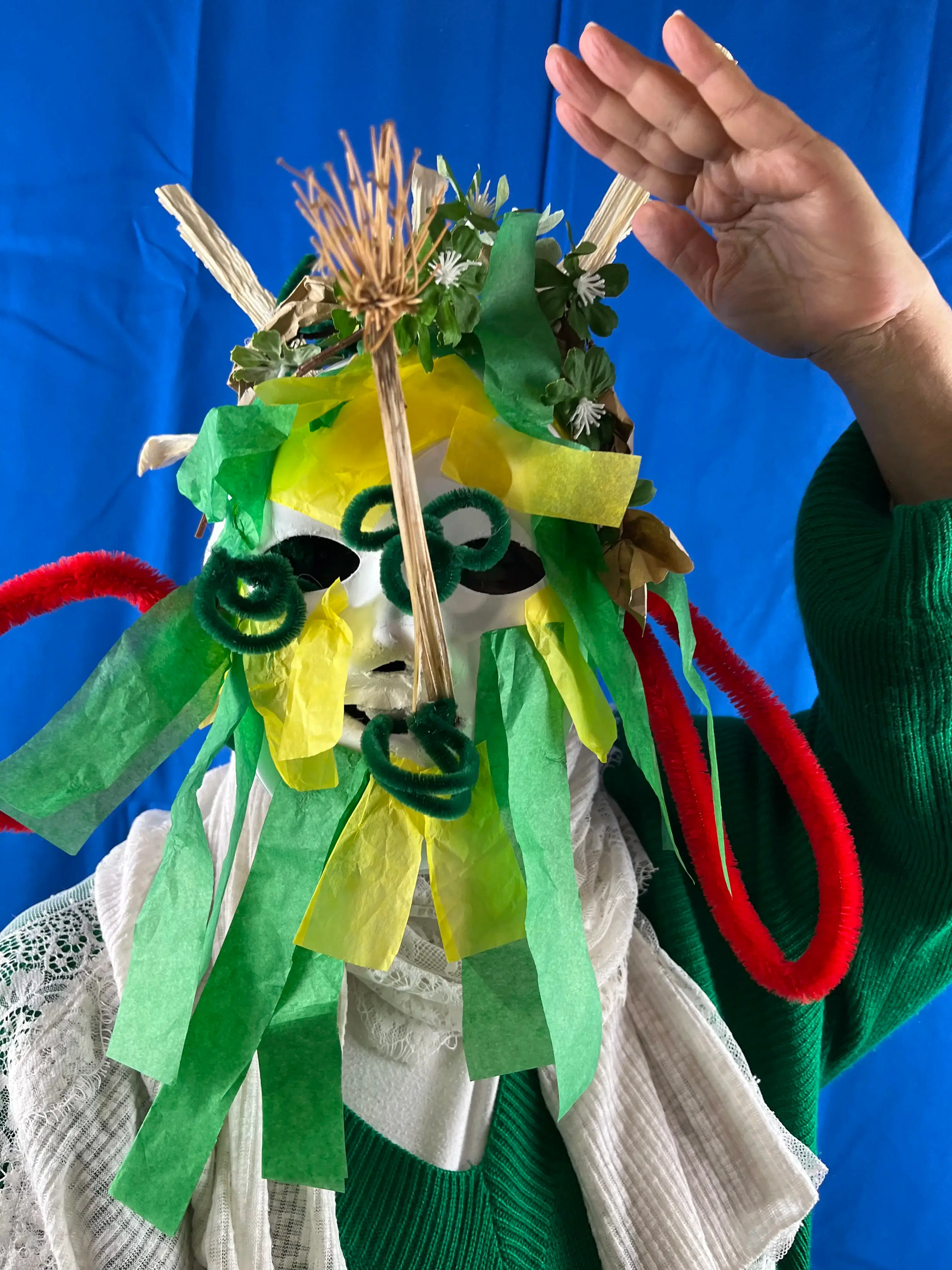 A vibrant handmade mask adorned with colorful paper, textured materials, and natural flora, held by a person against a blue backdrop.