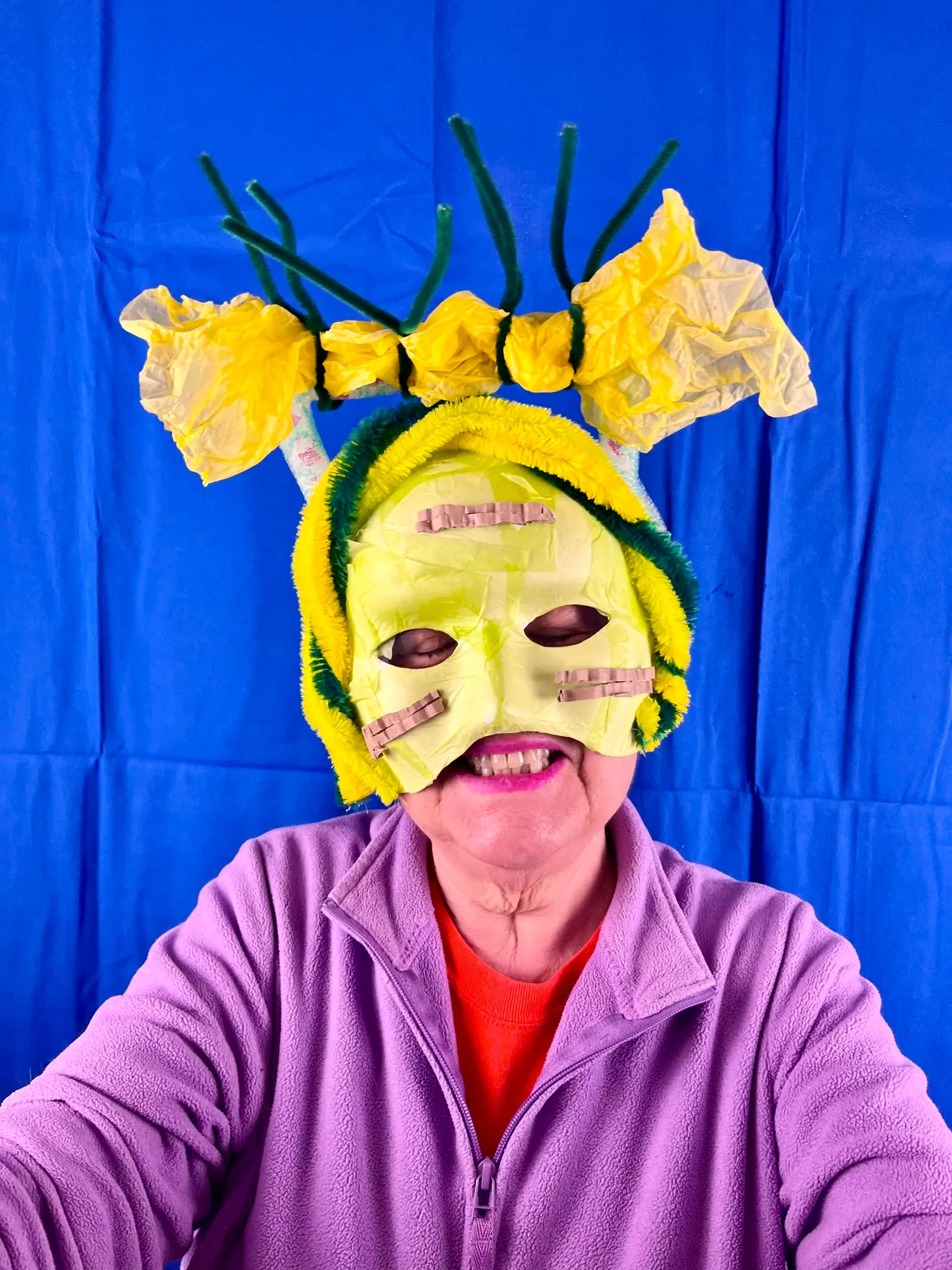 A person wearing a colorful handmade mask that resembles a whimsical creature, with yellow and green elements and protruding pipe cleaner antennae, against a bright blue backdrop.