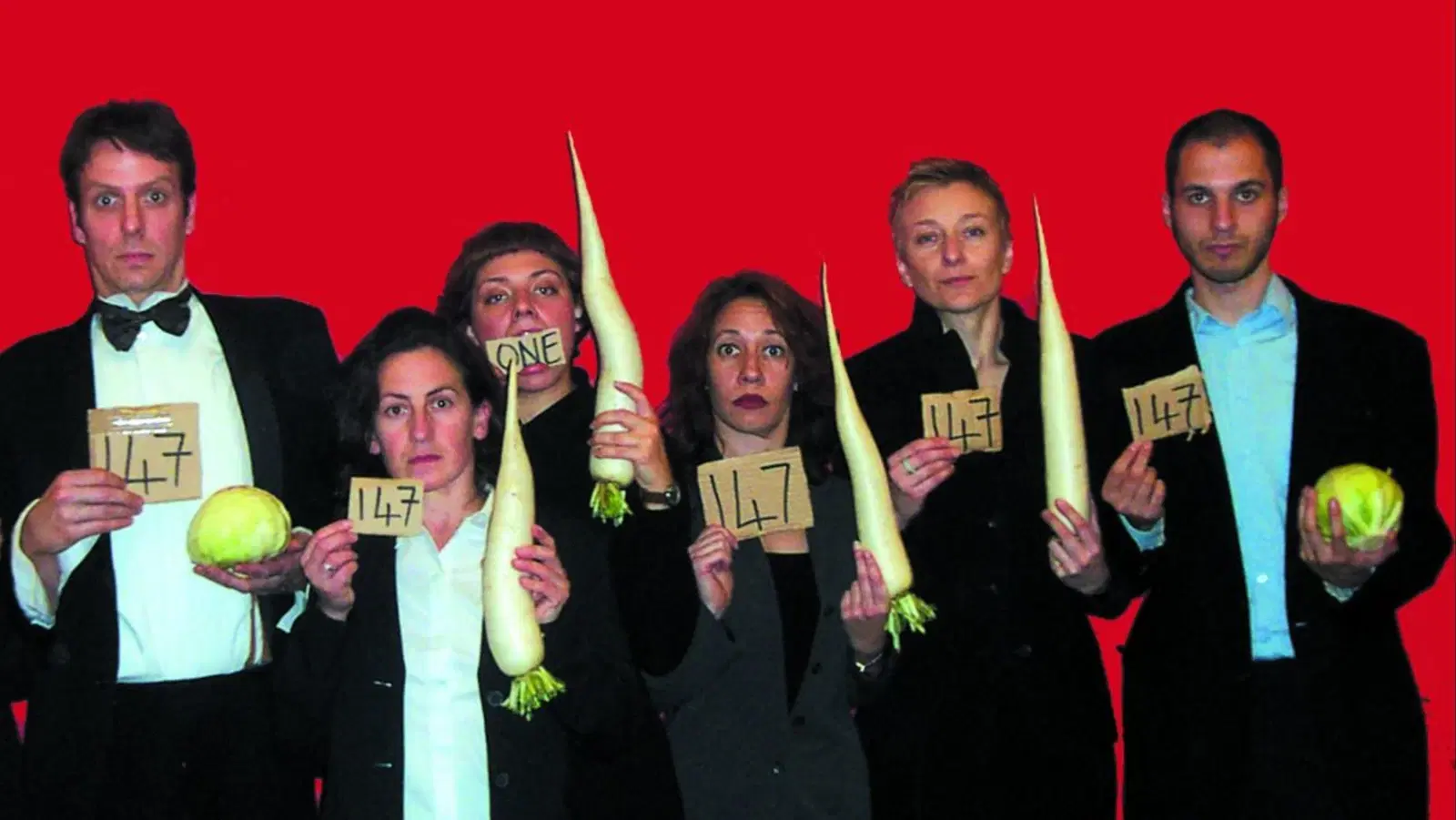 A group of six people holding vegetables and displaying numbers on cards, with puzzled expressions against a red background.