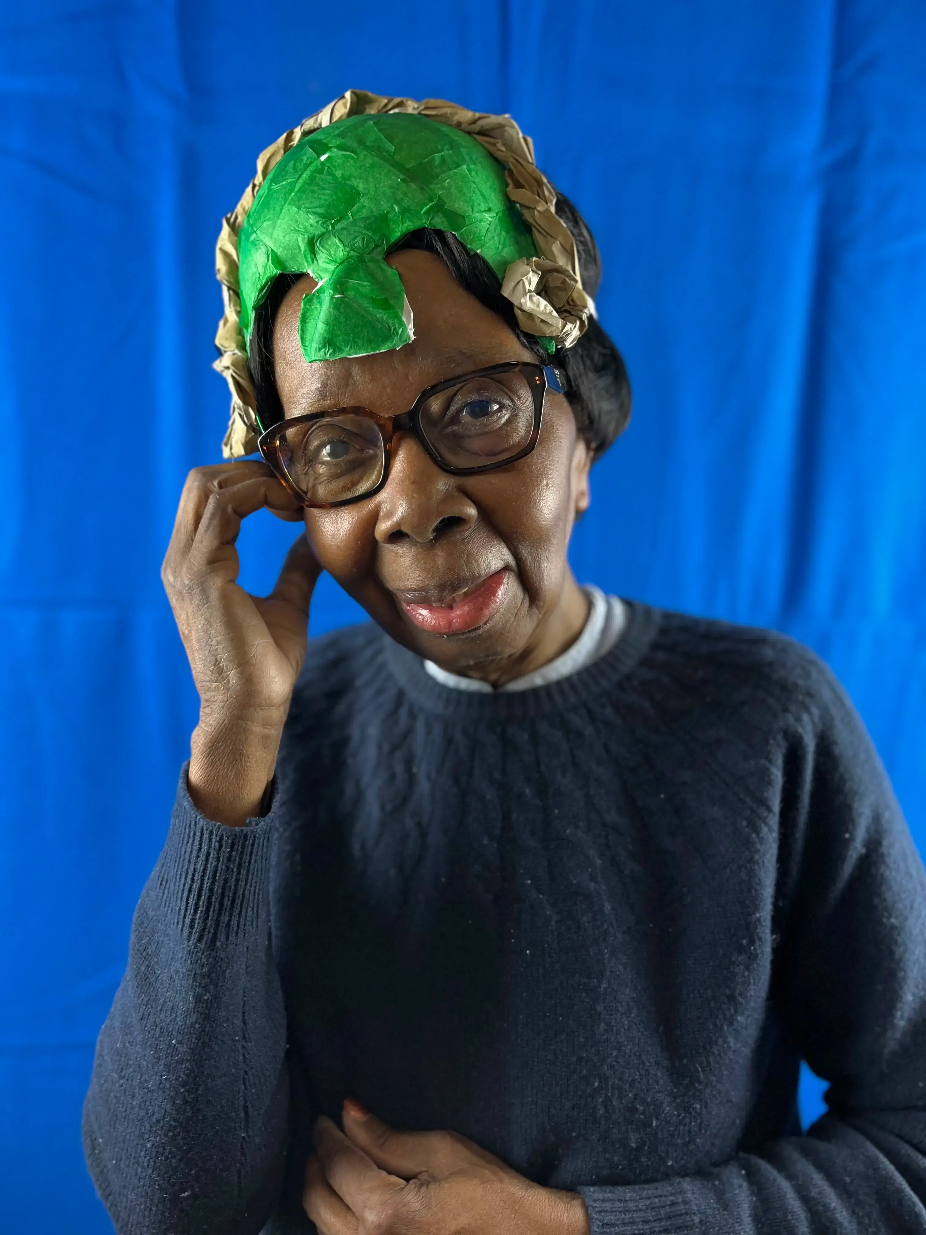 A portrait of a smiling woman wearing glasses and a colorful headwrap against a blue backdrop, with one hand playfully posed at her temple, exuding wisdom and vivacious spirit.