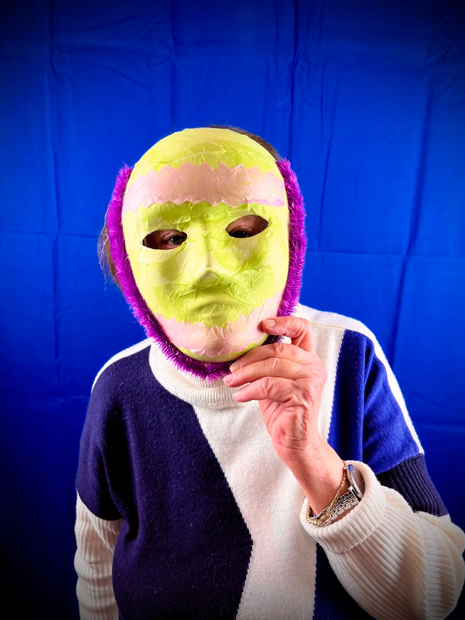 A person in a striped sweater standing against a blue background, wearing a colorful, abstract mask and looking directly at the camera.
