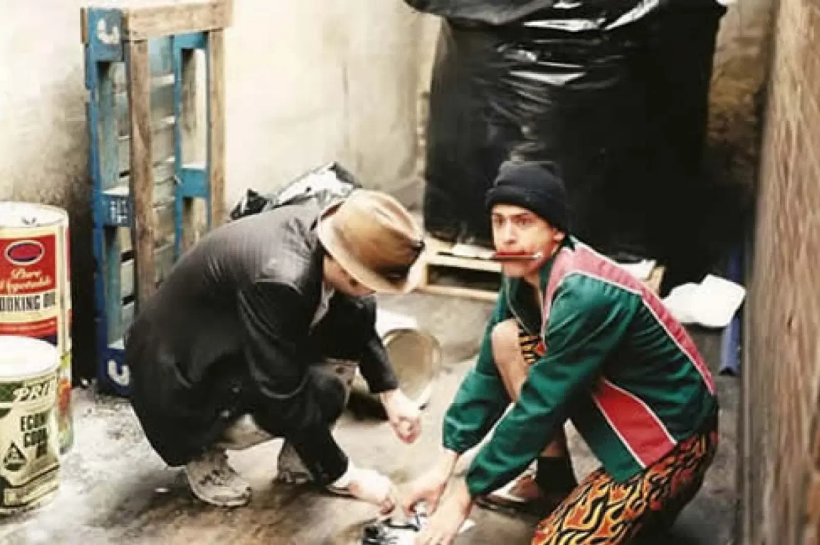 Two individuals squatting on a concrete floor engaged in an activity together, surrounded by various items and with a stark urban backdrop.