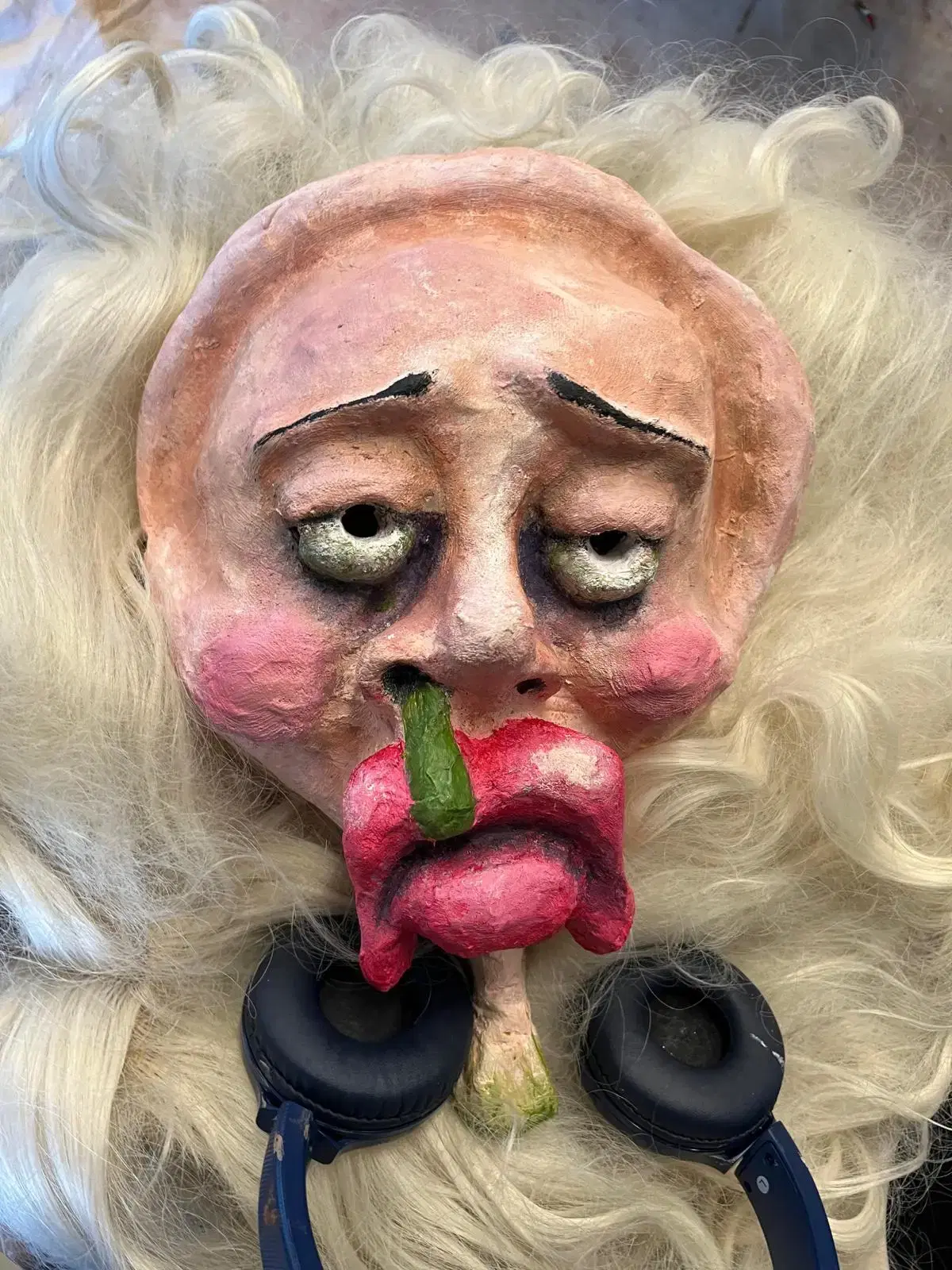 An image of a theatrical mask with exaggerated features, including a large red nose, puffy cheeks, and long white hair, comically and dramatically sticking out its tongue with a green chili pepper positioned on it. headphones rest around the neck of the mask, adding a modern touch to this otherwise classic representation of clown-like expression.