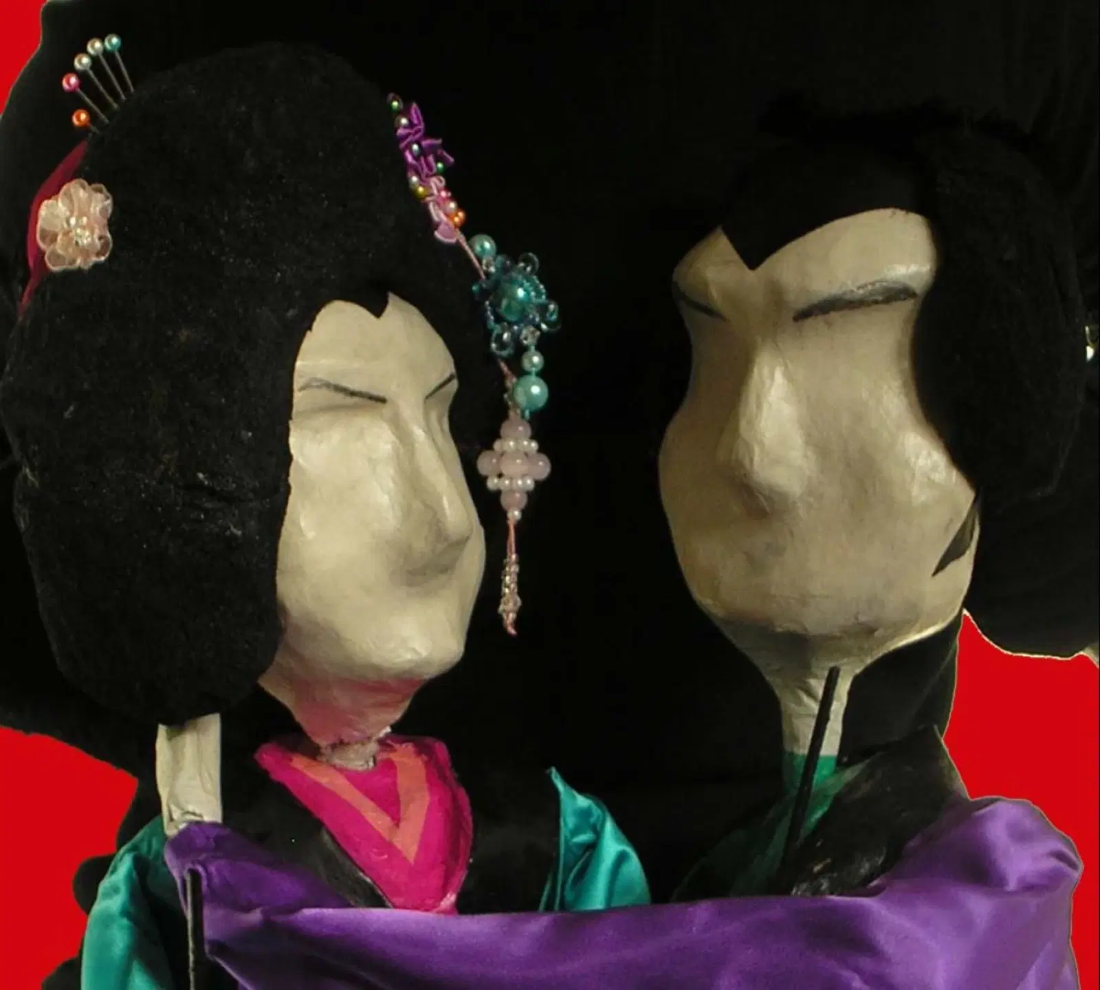 Two traditional asian-style puppets adorned with elaborate hairstyles and colorful costumes facing each other against a dark background.