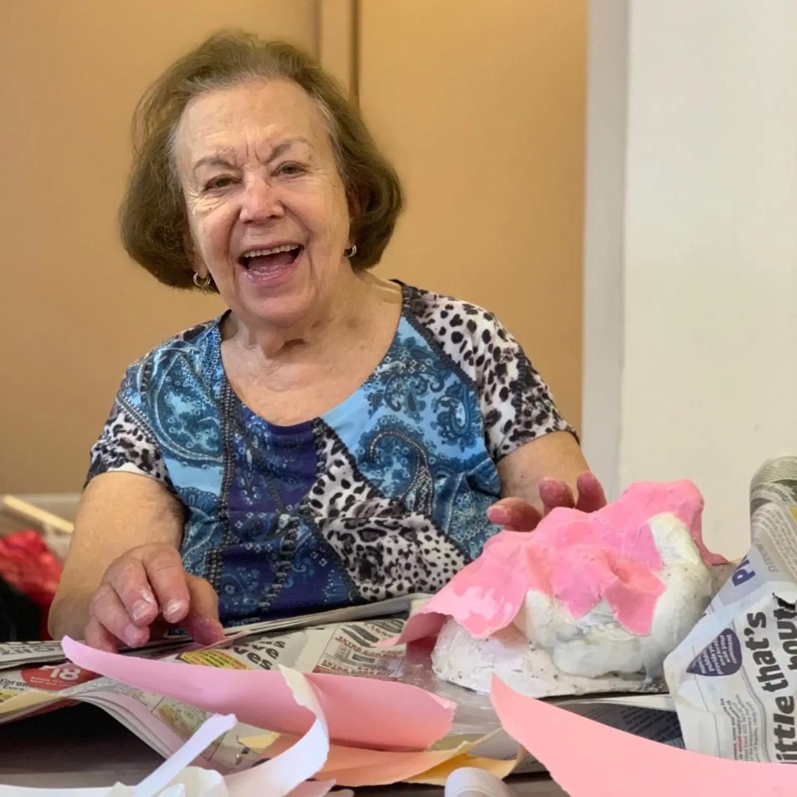 An elderly woman with a joyful expression sitting at a table engaged in a craft activity, with a colorful paper-mâché project in front of her.