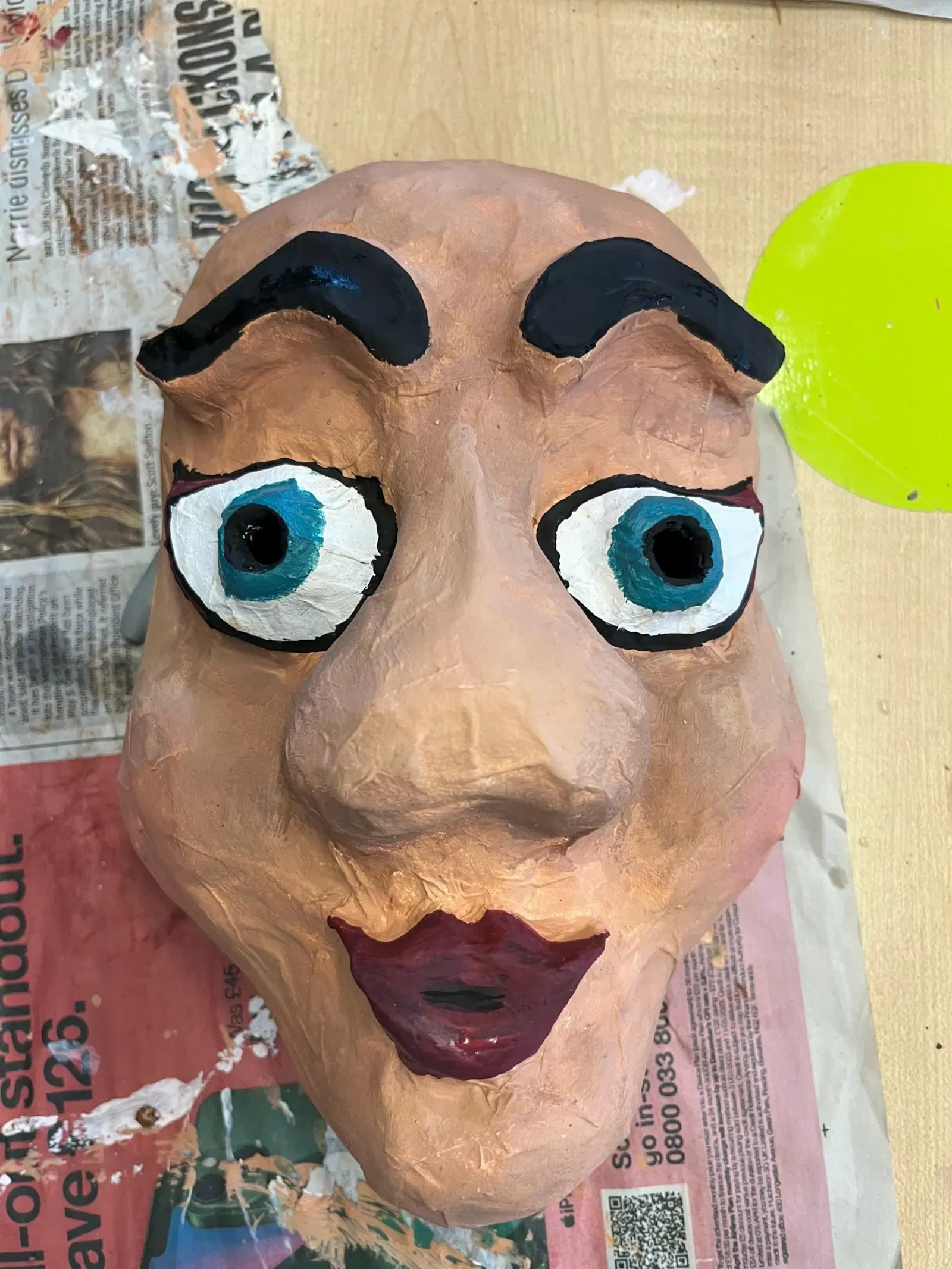 A handcrafted mask with exaggerated features including arched brows, wide eyes, and full lips, resting atop a printed paper surface.