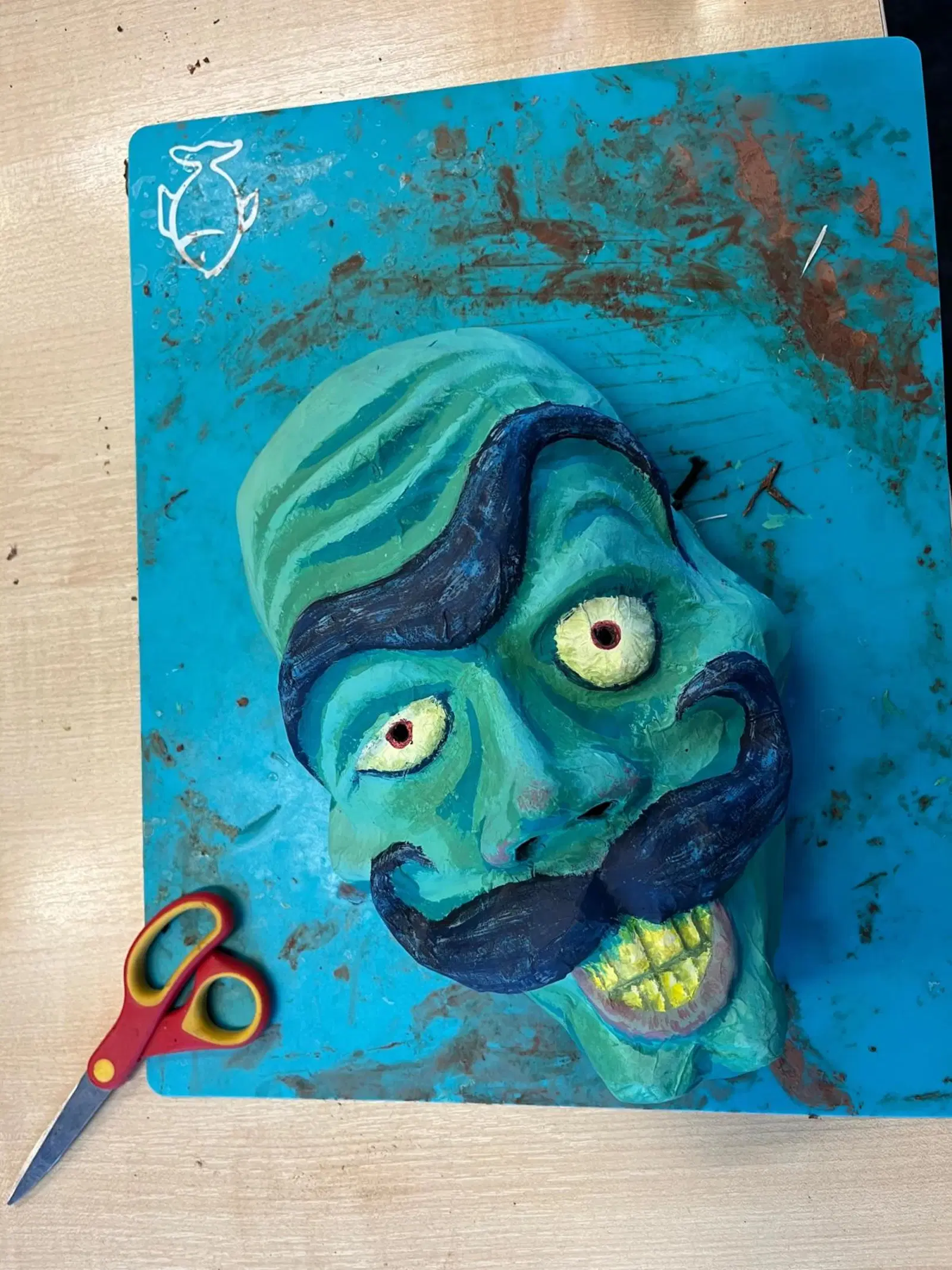 A vibrant clay sculpture of a caricatured face with exaggerated features, resting on a blue, paint-splattered workspace alongside a pair of scissors.