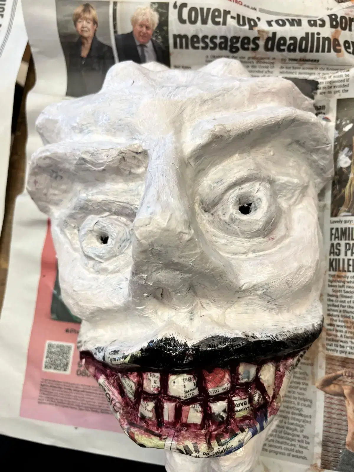 A close-up of a textured, painted clay or papier-mâché mask with exaggerated facial features, resting on an open newspaper.