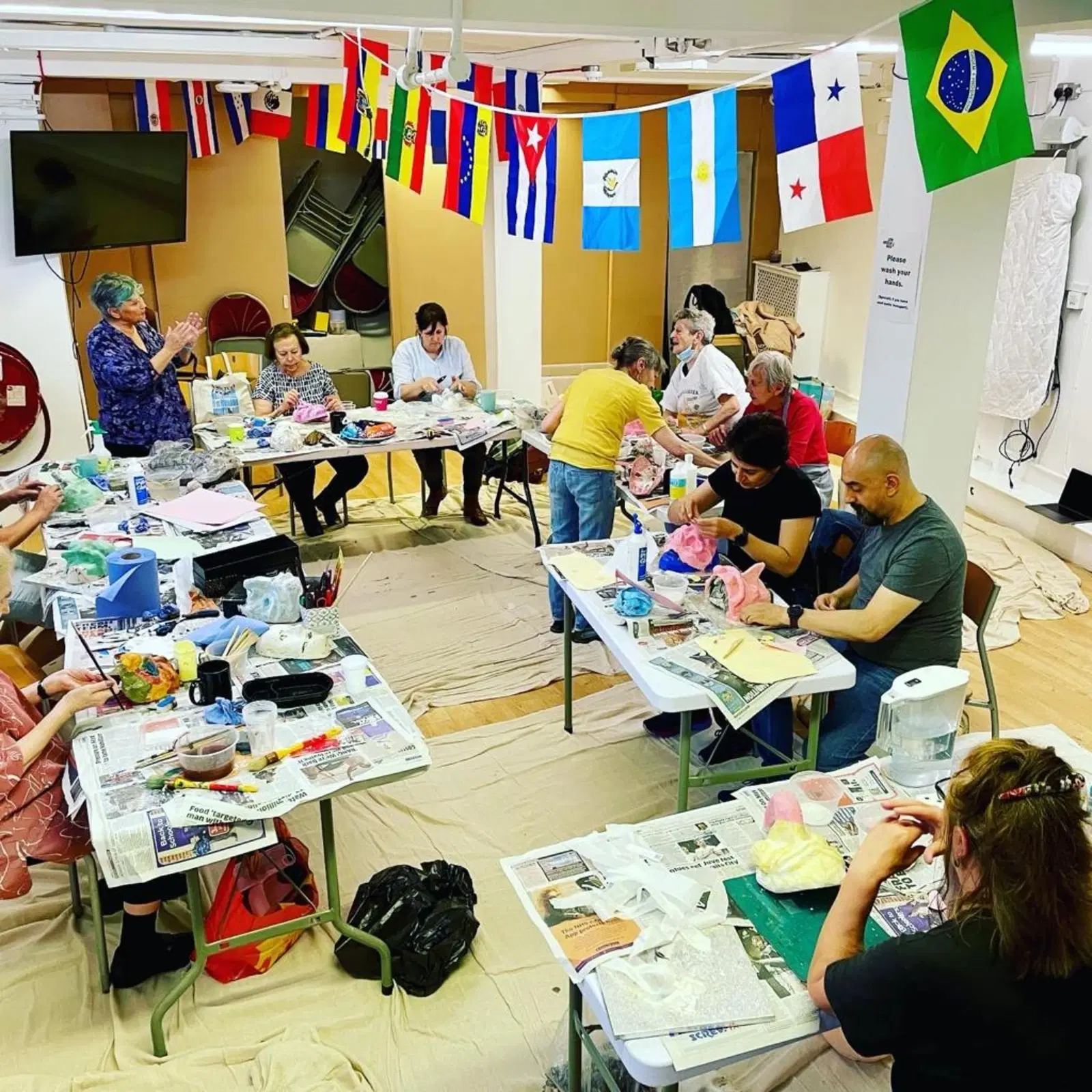 A vibrant art workshop in progress with participants of various ages focused on their craft, surrounded by colorful international flags hanging overhead, suggesting a culturally diverse or international event.