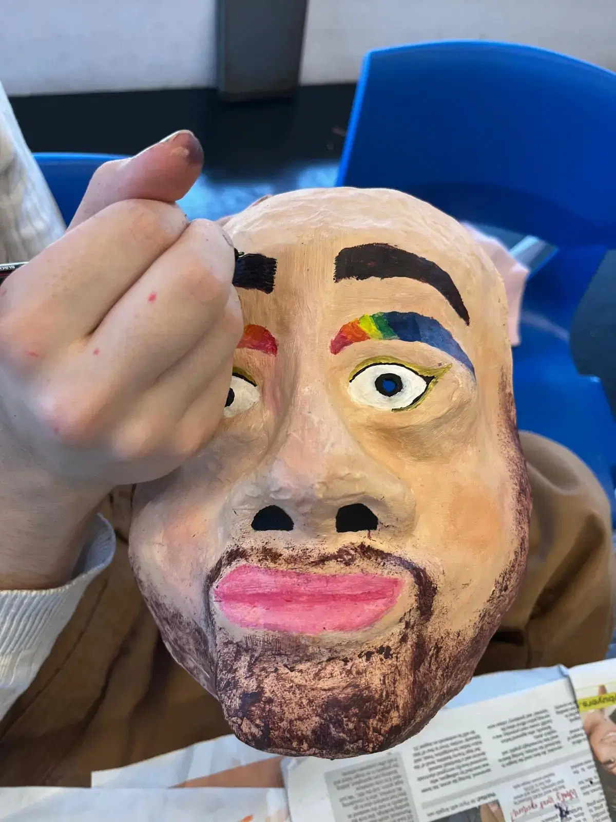 A hand holds up a handmade mask featuring a painted face with colorful eyebrows and pink lips, possibly a work-in-progress art project.